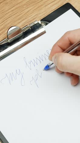close-up of a hand using a blue pen to write "my business plan" in cursive on a clipboard in vertical composition emphasizing focus and precision during early planning steps on a wood desk surface.