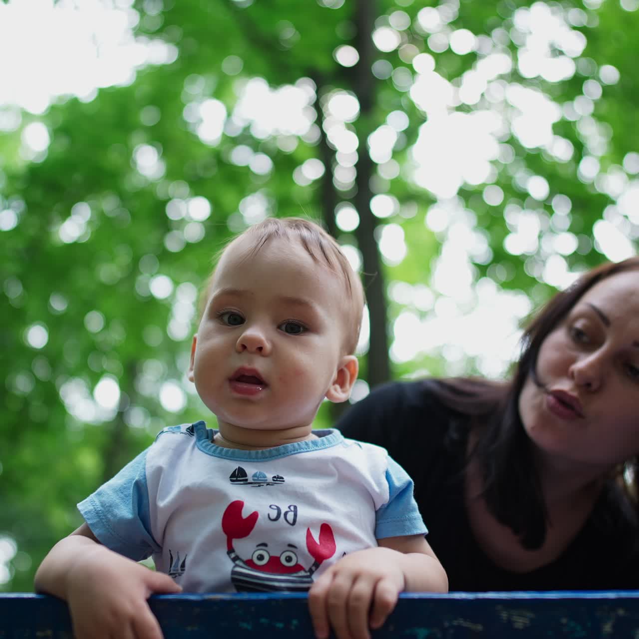 Curious lovely child outdoors. Baby boy standing on the bench looking at camera. Mother's behind her son. Low angle view