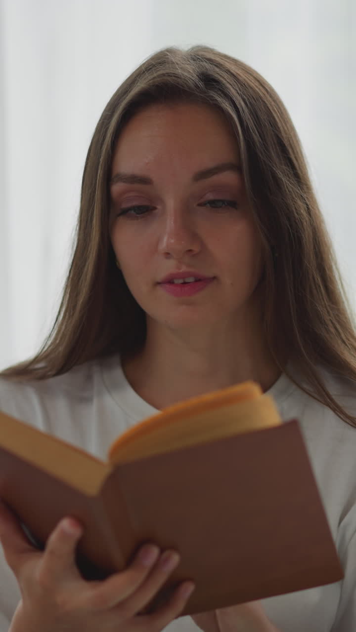 mujer enfocada se sienta en silla de ruedas leyendo un libro interesante contra una ventana brillante con cortina blanca. la dama sonríe disfrutando de la historia en primer plano