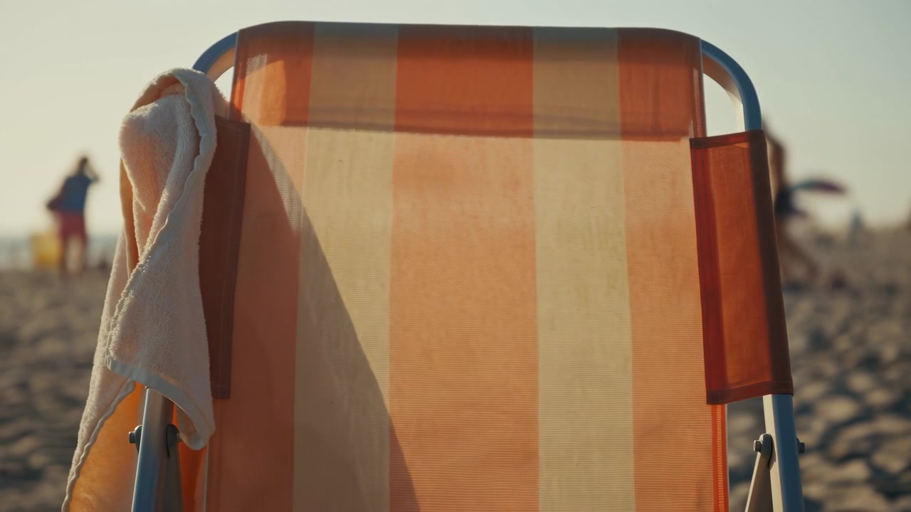 Vista frontale ravvicinata di una sedia a sdraio a righe arancioni e beige con un asciugamano di spugna bianco drappeggiato sul braccio sinistro, posizionata su una spiaggia sabbiosa illuminata dal sole in una calda luce dorata pomeridiana con uno sfondo costiero sfocato e morbido