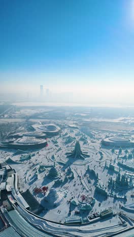 vertical aerial view of the harbin ice festival 2025, showcasing intricate ice sculptures, frozen palaces, and a vast winter wonderland set against the city skyline under clear blue skies. china