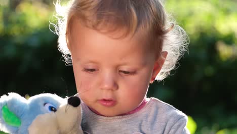 a little girl with blond curly hair sits in the yard of the house and gently hugs a teddy bear a little girl with blond curly hair sits in the yard of the house in sunny summer weather and gently hugs a teddy bear