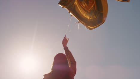 happy young woman celebrating birthday party holding golden balloons floating on sunny rooftop at sunset