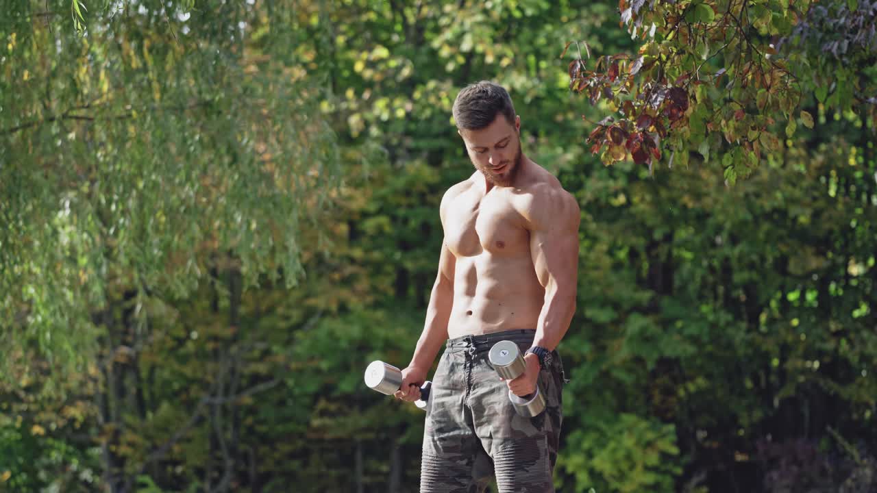 Bearded shirtless man doing workout in nature. Muscular bodybuilder exercising with dumbbells outdoors. Handsome athlete lifting dumbbells.