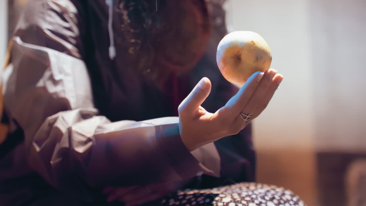Blødt indendørs nærbillede af hånd, der balancerer en appelsin, siddende kvinde justerer frugten med fingerspidserne, mønstret nederdel og jakke, varm vinduesglød, oprigtig bevægelsesstudie, blidt kast og fangst, kontemplativ.
