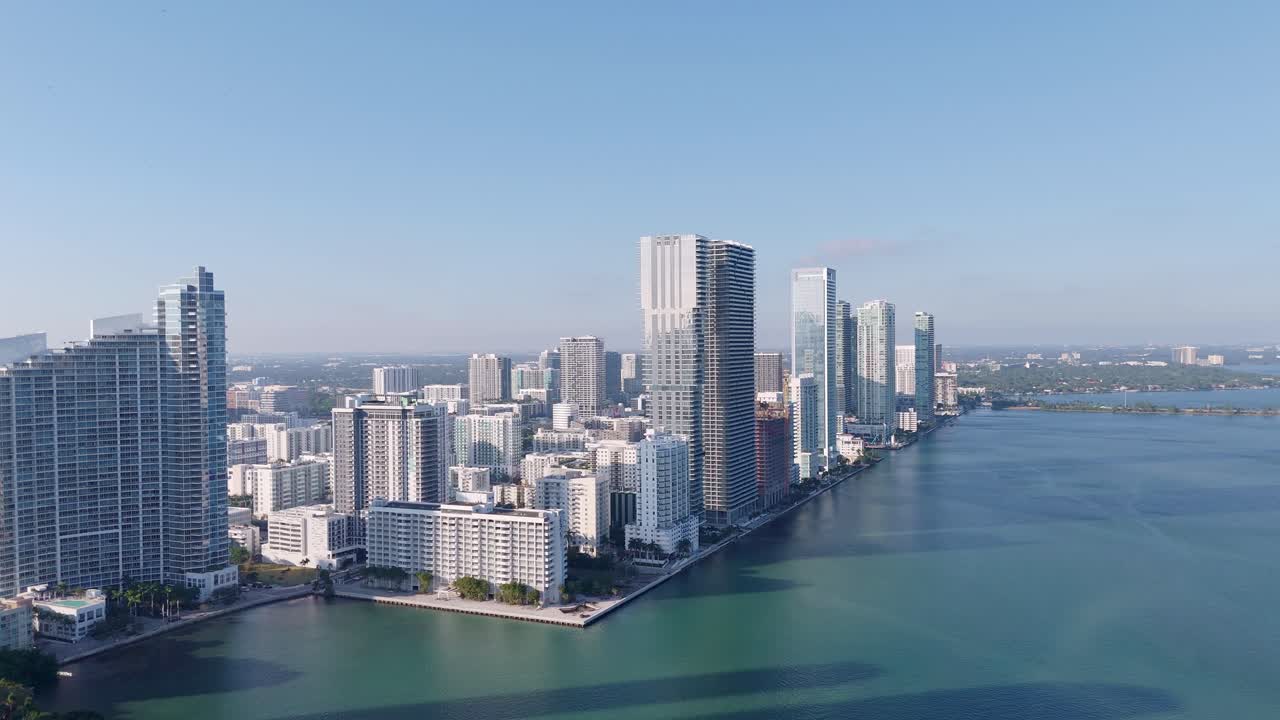 Aerial drone view of Miami’s coastal skyline with modern high-rise buildings along Biscayne Bay, showcasing turquoise waters and a vibrant urban landscape under clear blue skies.