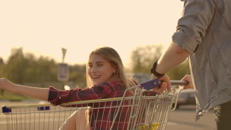 Cupé-Joven-Y-Elegante-Divirtiéndose-Montando-Con-Un-Carrito-De-Compras-En-El-Estacionamiento-Al-Aire-Libre-Cerca-Del-Supermercado