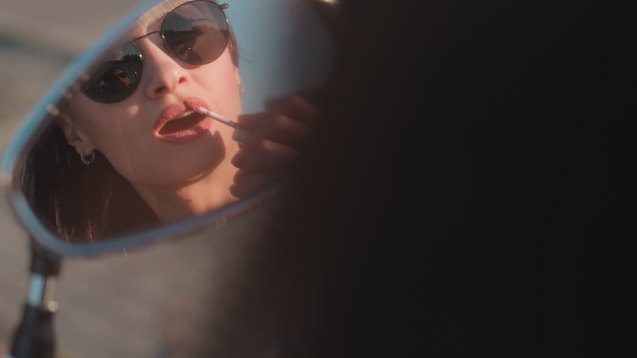 Lo specchietto laterale di una moto riflette una donna elegante con occhiali da sole che applica attentamente il rossetto alla luce del sole, mettendo in risalto bellezza, sicurezza ed eleganza con una prospettiva creativa