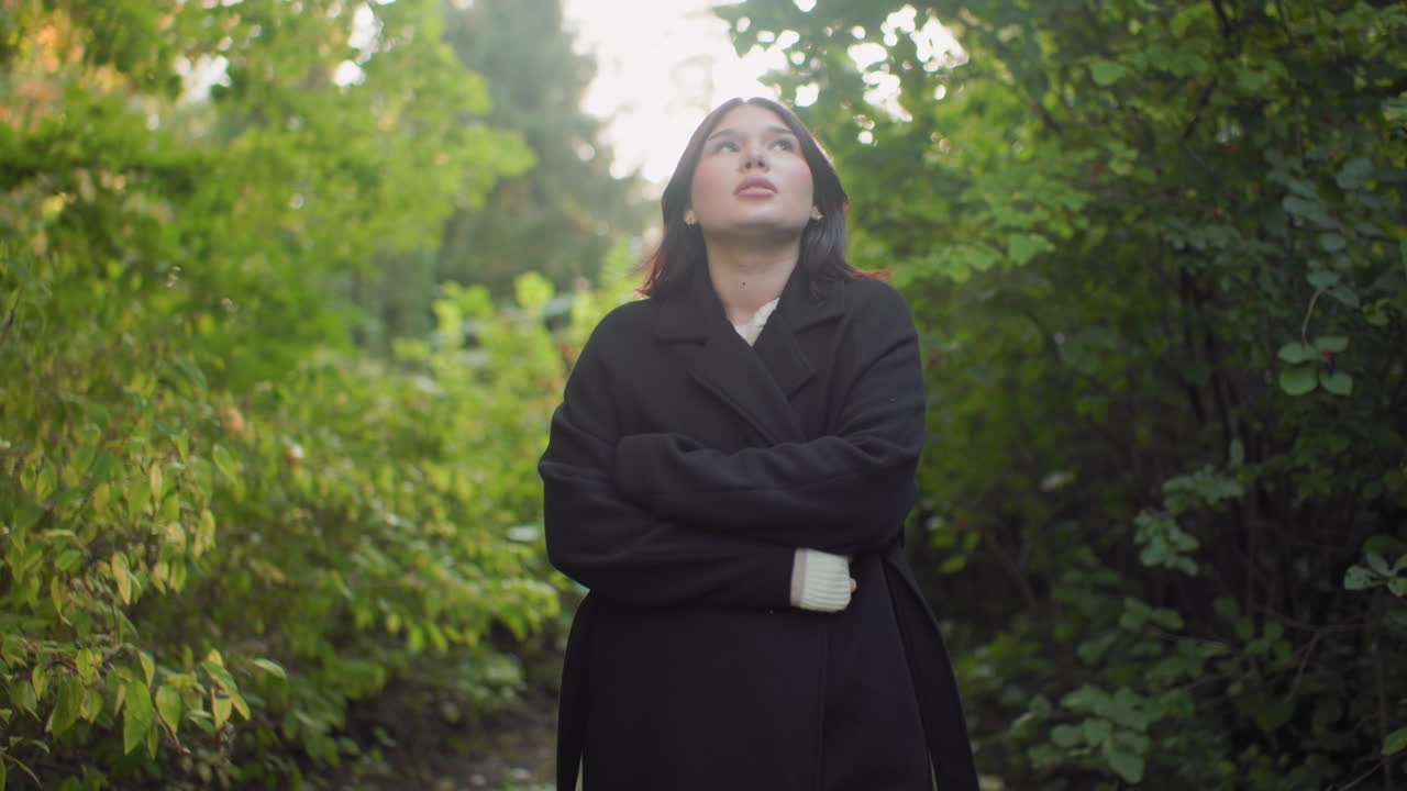 Cozy young lady adjusting jacket while walking through green forest path, looking around with calm curious expression, peaceful stroll in quiet park creating warm seasonal mood