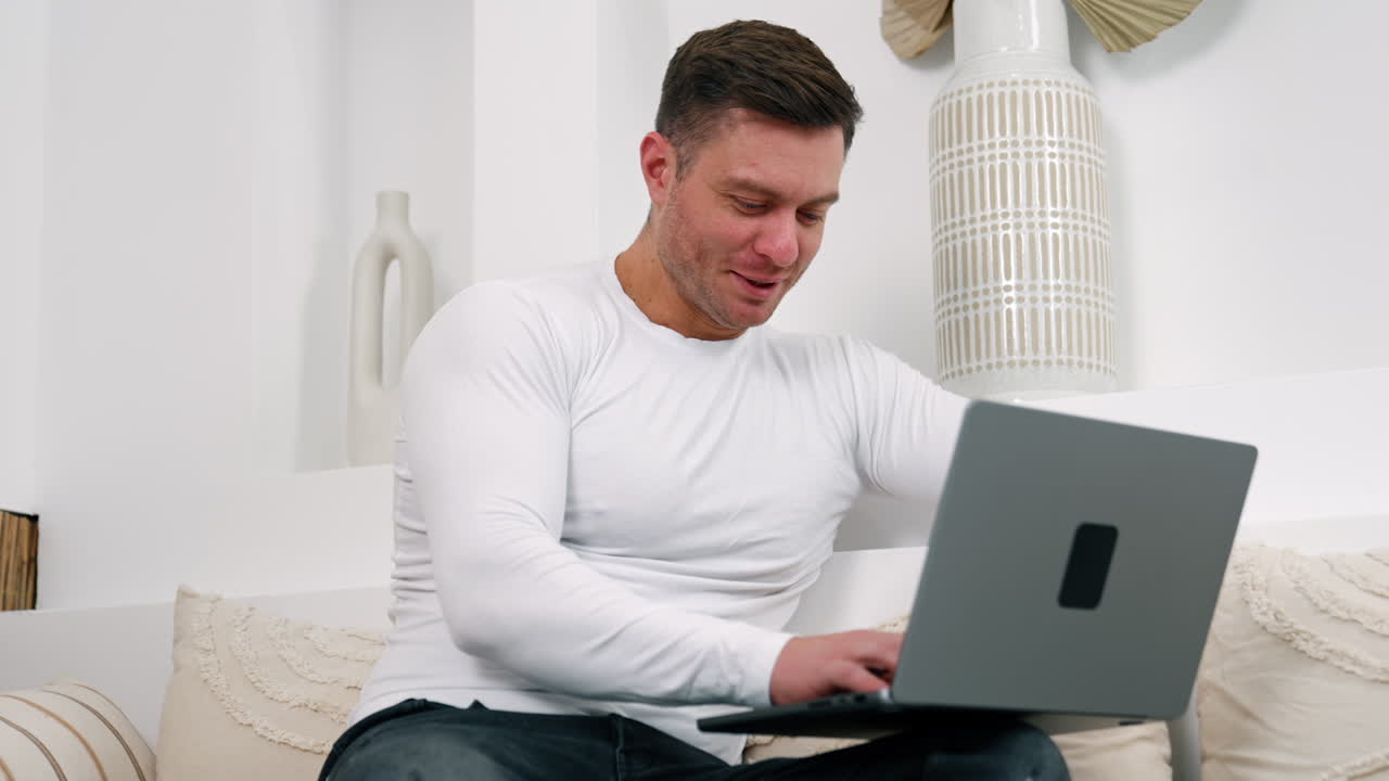 Attractive young man in white sweater typing on his computer. Caucasian male sits on sofa working comfortably from home.