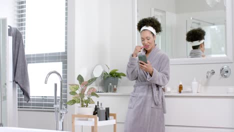 mujer biracial en bata de baño cepillándose los dientes y usando un teléfono inteligente en el baño, cámara lenta