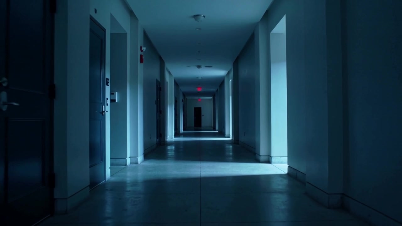 Carrellata che rivela un corridoio d'ufficio vuoto con luce a pavimento e insegna di uscita rossa, evidenziando il vuoto.