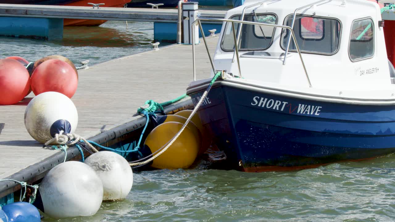 Small blue and white fishing boat rocks among buoys in a bright, choppy harbor scene