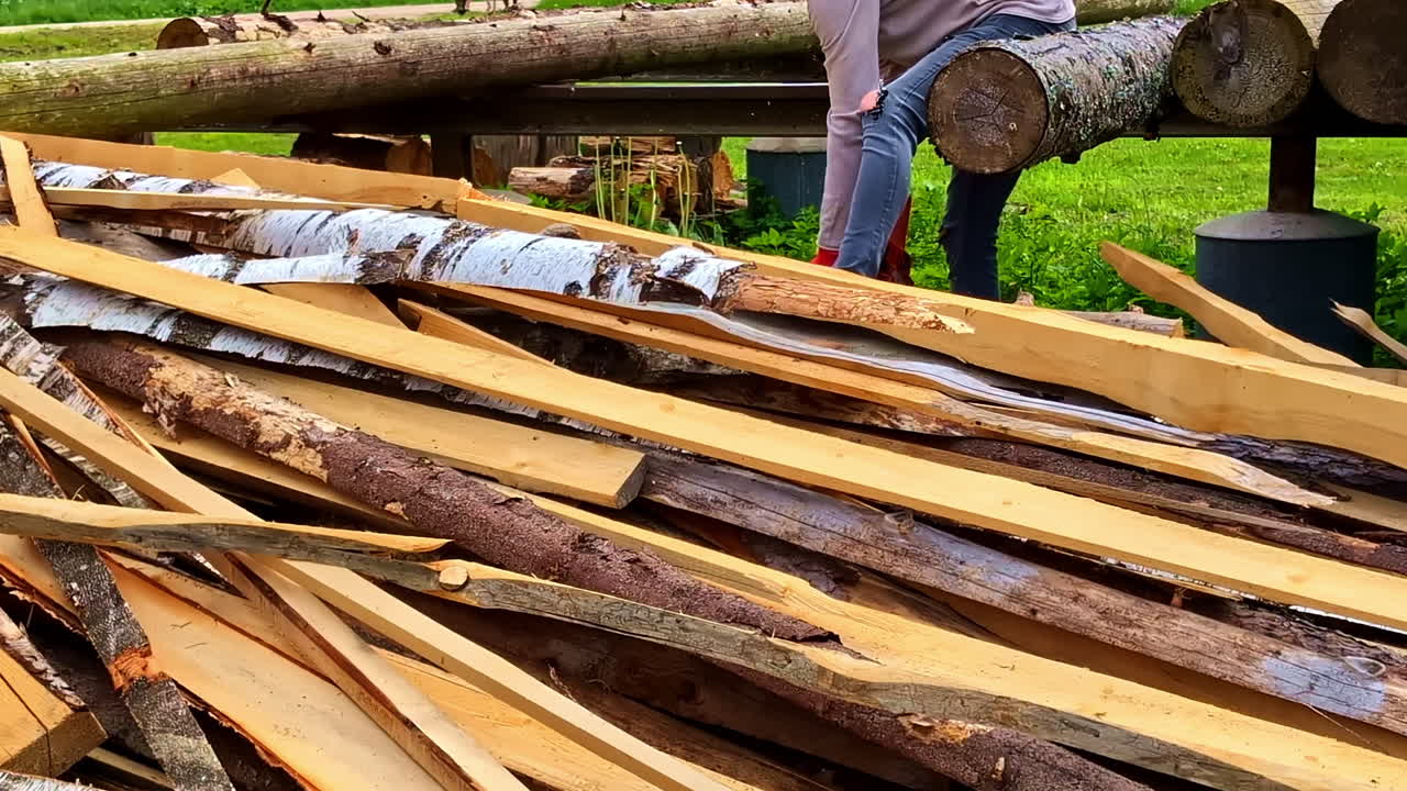Man Using Chainsaw to Cut Wooden Planks Beside Timber Pile Outdoors