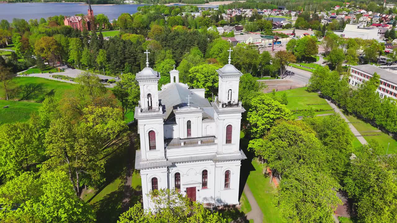 Aerial footage of a white twin-tower church nestled in green spring foliage, with surrounding paths, homes, and river in a picturesque townscape.