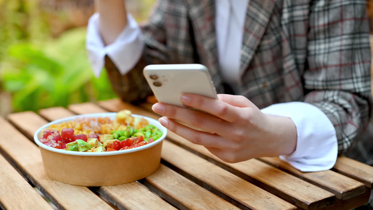 View of a young woman eating poke bowl with vegetables and fruits using chopsticks while sitting in smartphone. Park with a lot of greenery and illumination. Slow motion