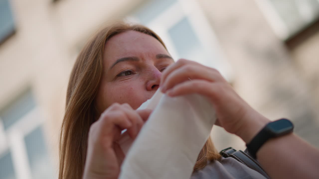 Sad mum standing outdoors near blurred building background cleaning tears with wipes, emotional face showing grief and quiet sorrow, hair moving in soft wind