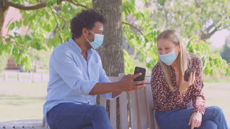 socially distanced couple in masks meet in park during health pandemic looking at mobile phones