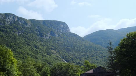 granite mountains covered in vegetation tower over treetops as the camera pans from a low angle against a blue sky