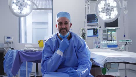 Portrait-of-caucasian-male-surgeon-wearing-lowered-face-mask-sitting-in-operating-theatre-smiling