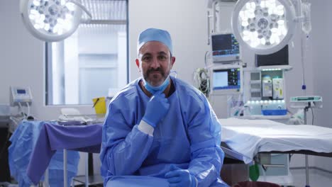 retrato de un cirujano caucásico con una máscara facial baja sentado en la sala de operaciones sonriendo