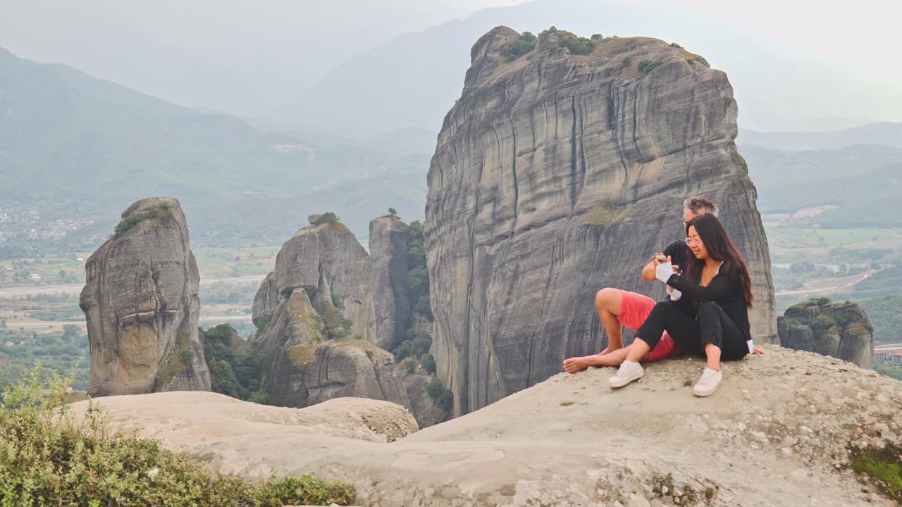 Romantic couple take selfies sat on mountain overlook Meteora landscape