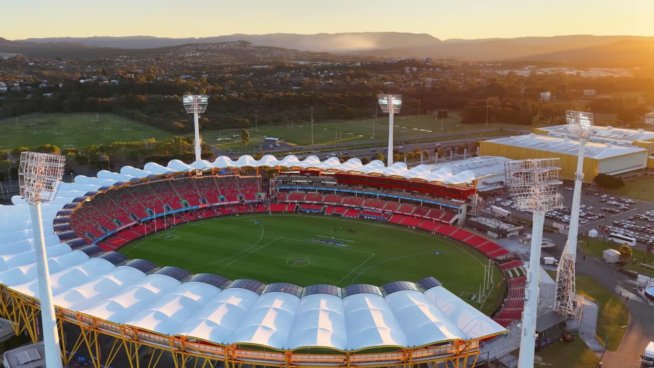 Drone footage glides over a large illuminated stadium at sunset, revealing the venue, green field, parking lots, and scenic hinterland landscape in golden light