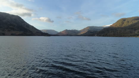 low aerial drone shot flying over ullswater lake at sunrise with helvellyn mountain in background on sunny morning with clouds lake district cumbria united kingdom