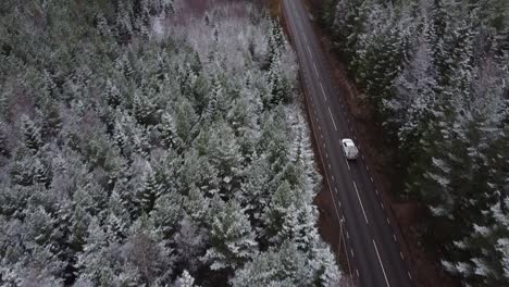 slow moving drone shot of a car driving up a road and driving through frosty forest and paved road