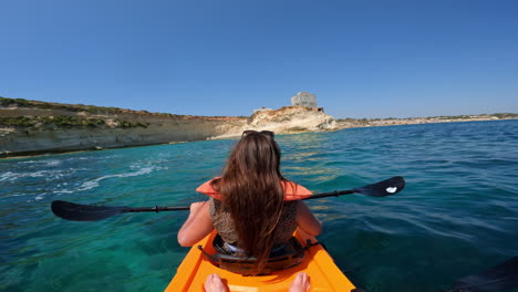A-woman-in-an-orange-kayak-rowing-towards-the-rocky-coast-of-Malta