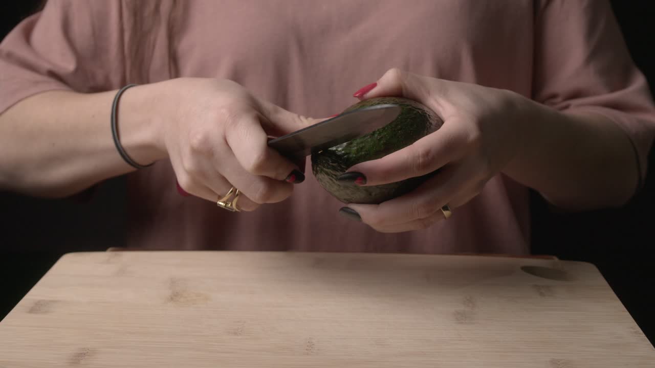 In a dimly lit scene, a woman’s hands skillfully cut open a ripe avocado with a knife, set against a dark background