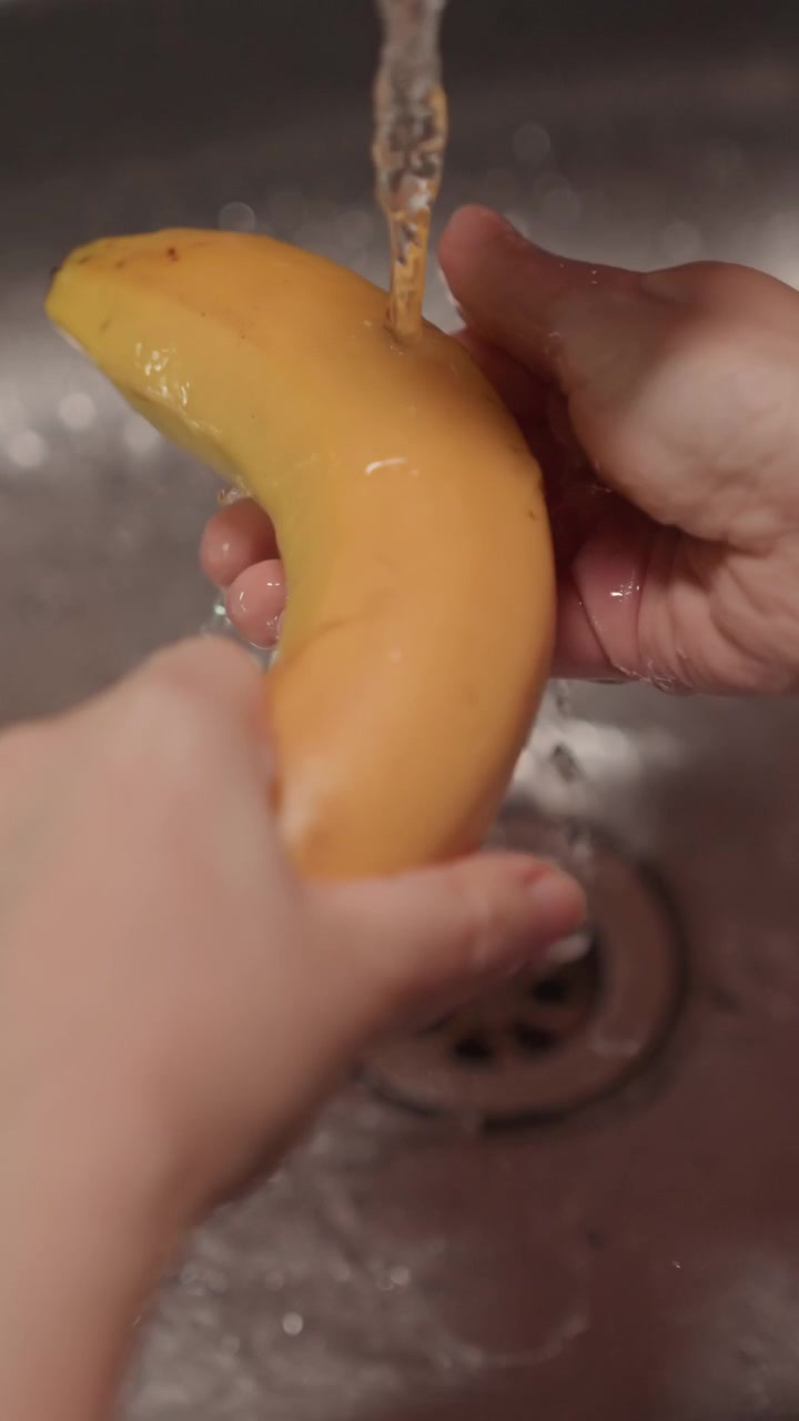 Primer plano de manos lavando un plátano amarillo bajo el grifo en el fregadero. La fruta se gira y se frota para limpiar todos los lados. Fondo de cocina de acero inoxidable con enfoque en el desagüe del grifo
