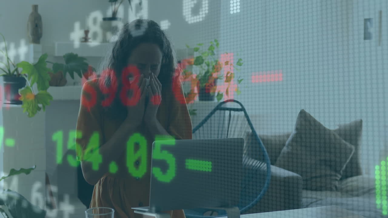 Mid-adult woman holding tissue and watching laptop screen at desk, with red green stock charts