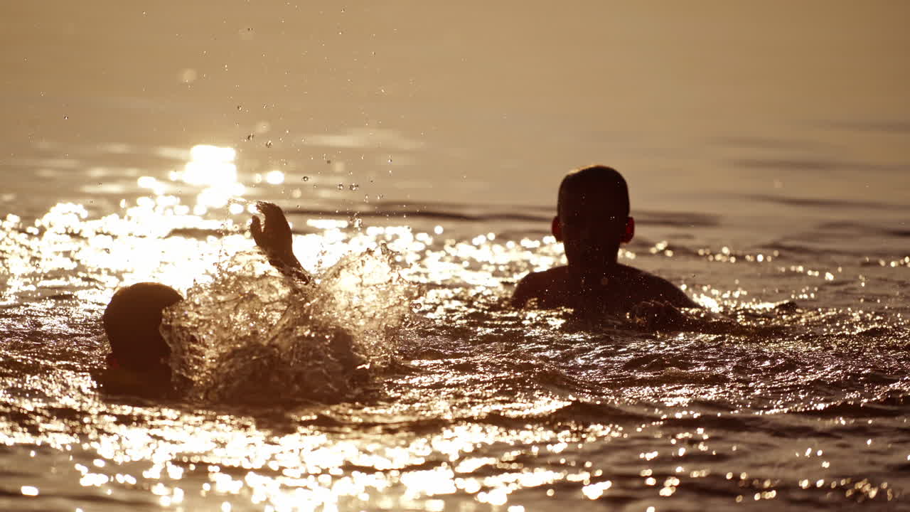Two boys splashing with water happily at sunset. Silhouette of kids heads playing in the river water during summer holidays in the evening. Joyful summertime.