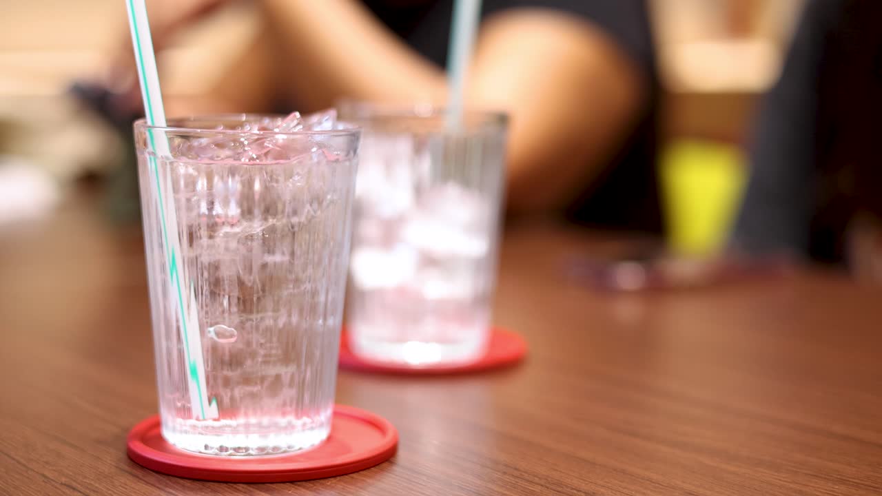 A person’s hand approaches, lifts, and sets down a glass of iced water with a straw on a red coaster in a warmly lit indoor setting