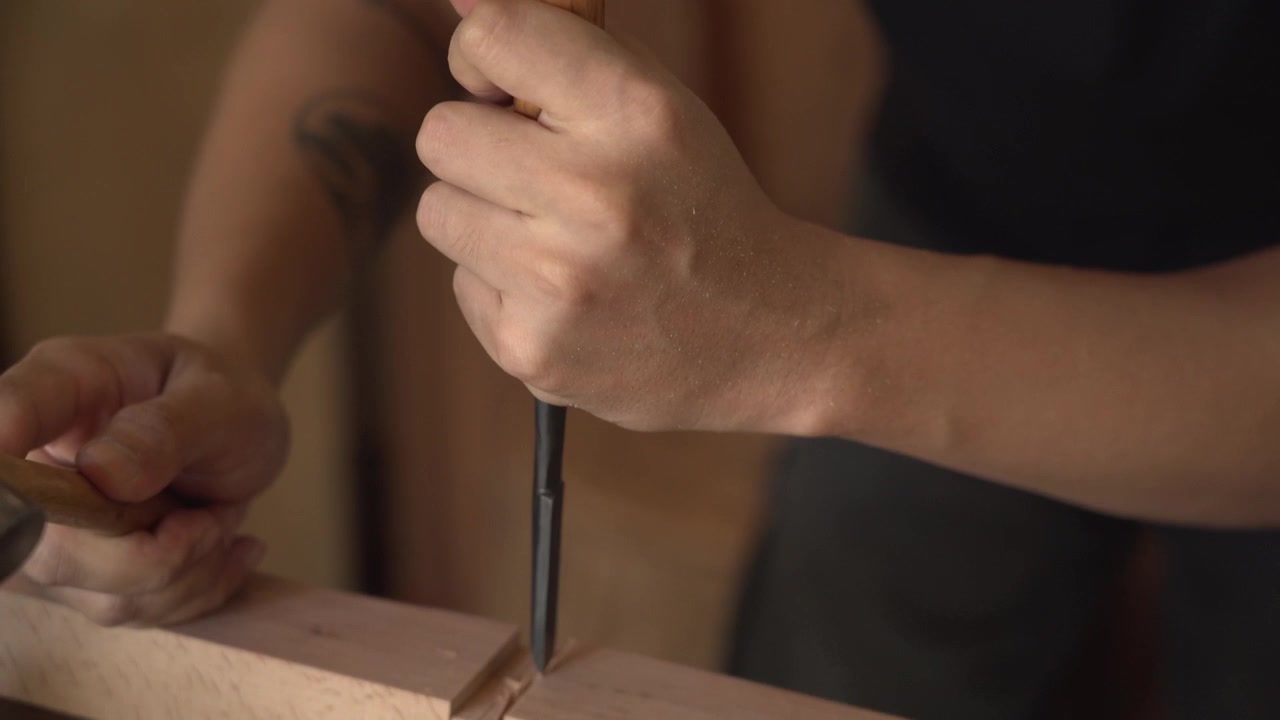Close up of craftsman chiseling groove in wooden beam with mallet and chisel, removing wood to form joinery during woodworking process on workbench