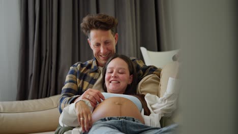 happy middle aged man in a plaid shirt stroking the belly of his pregnant wife while sitting on the sofa at home while relaxing