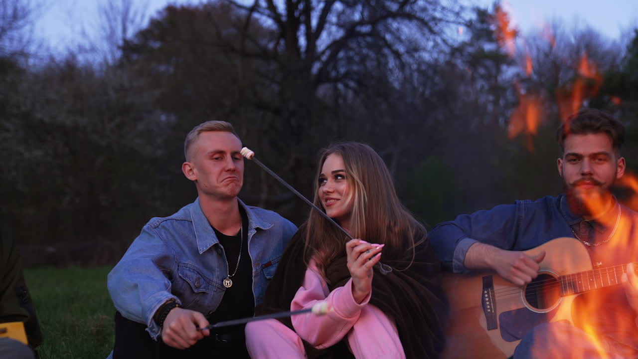 Dating couple making marshmallows on fire. Friends nearby playing music and singing songs. Nice summer evening in the nature.