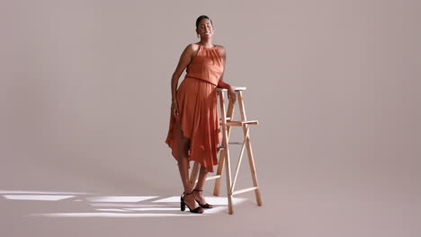 indian female model entering frame posing on ladder in sunlit studio showing burnt orange dress