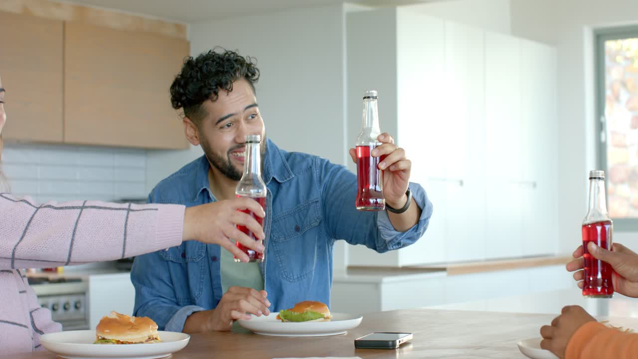 Brinde motivacional de amigos diversos batendo as garrafas na ilha da cozinha enquanto compartilham hambúrgueres e refrigerante