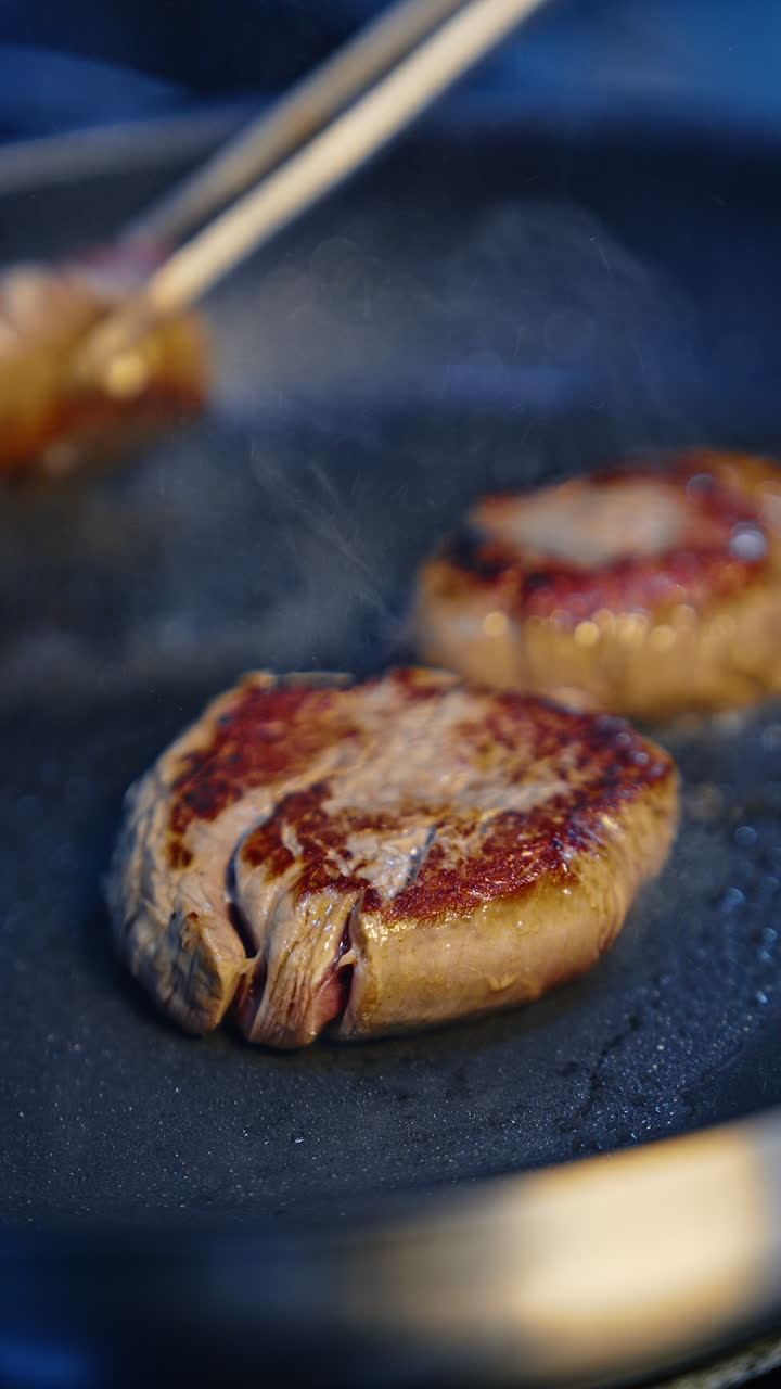 Metal tongs turn the chops of medallion steaks fried on the frying pan. Cooking red meat. Close up. Vertical video.