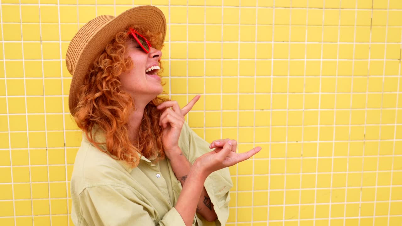 Woman in hat and sunglasses posing against yellow tiled wall