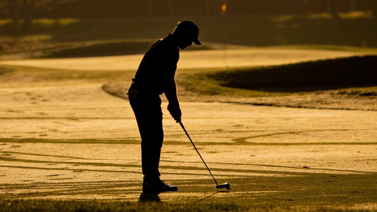 Golfista con cappellino e scarpe si posiziona sulla palla, stringendo la presa sul putter su un green illuminato dal sole, preparandosi al putt.