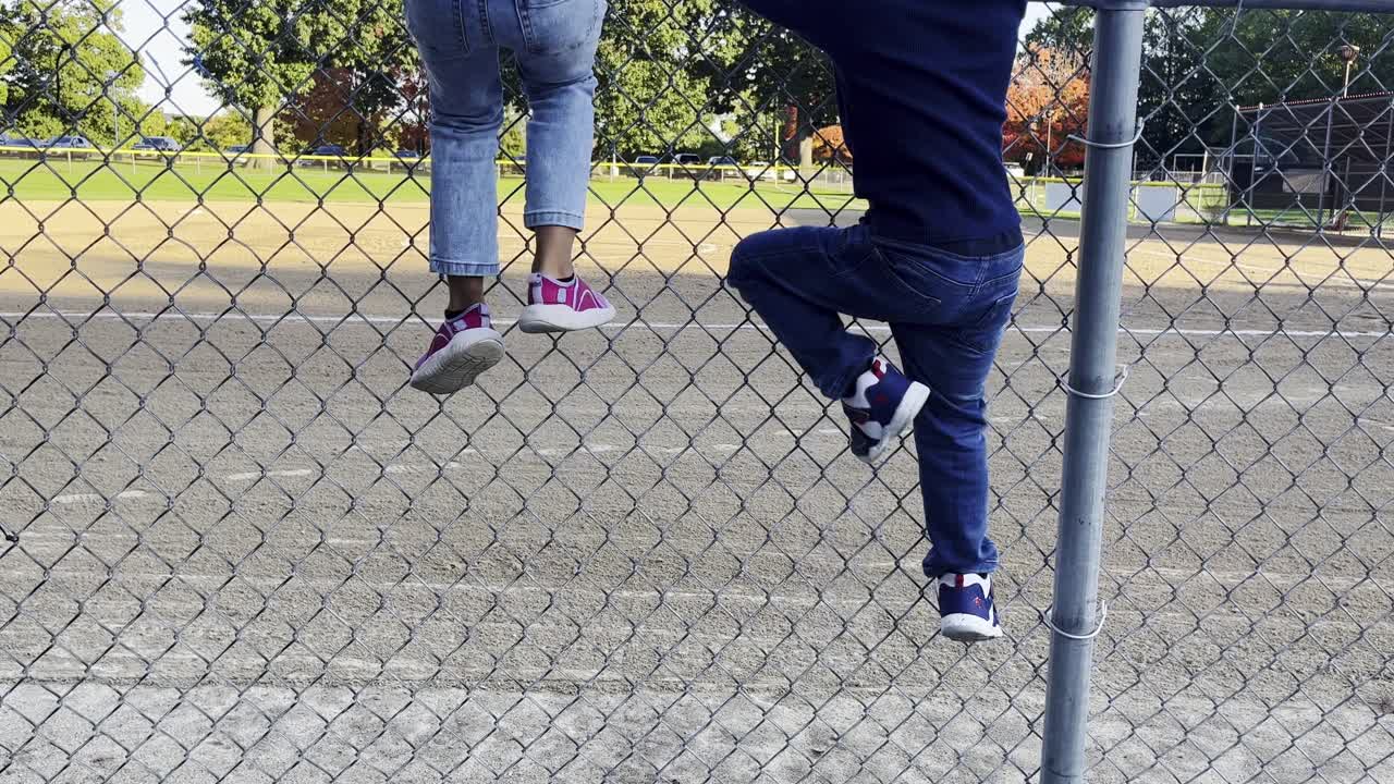 dos niños pequeños escalando una cerca de eslabones de cadena en el campo de softbol en un día soleado, cierren la toma estática