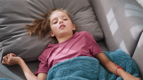 restless girl lying on couch, covered with soft blue blanket, looking unwell and sick, showing discomfort, indicating cold or flu symptoms with tired expression