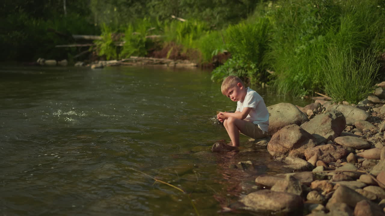 Serious boy sits on bank rock contemplating breathtaking view in wild nature park on sunny day. Cute child looks at shallow river enjoying scenery and fresh air