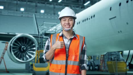 ingeniero masculino asiático con casco de seguridad de pie con aviones en el hangar. sonriendo y mostrando los pulgares hacia arriba gesto a la cámara mientras el mantenimiento de aviones