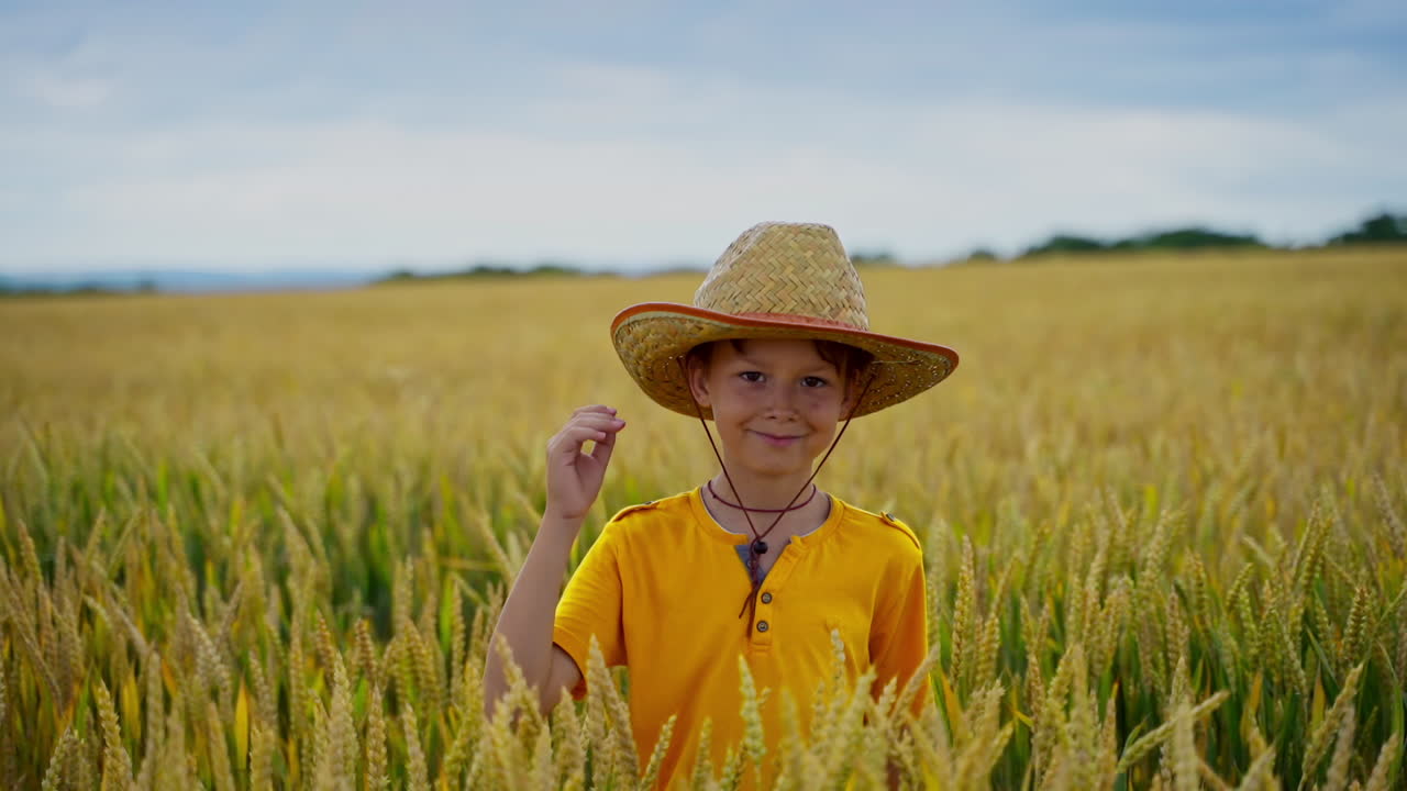 Portrait of little agronomist. Cute boy in yellow t-shirt and straw hat standing among wheat field in summer day. Smiling kid in agricultural land.