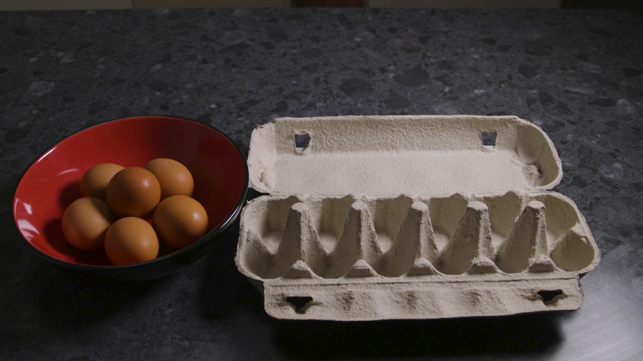 Braune Eier bewegen sich in einer Stop-Motion-Sequenz aus einem Eierkarton in eine rote Schüssel auf einer dunklen Küchenarbeitsplatte.