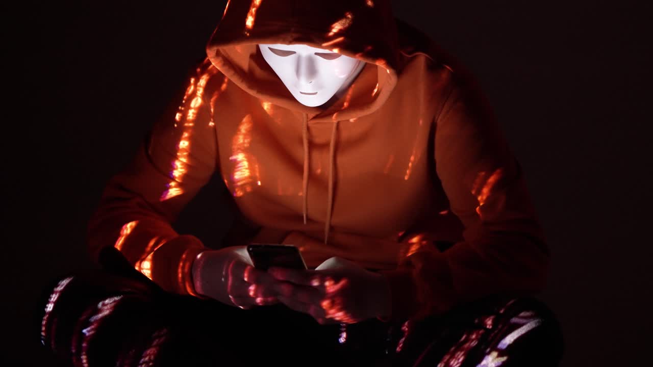 A faceless hacker in an orange hoodie is illuminated by red code reflections while using a smartphone in the dark, highlighting covert digital activity and mobile-based cyber interference